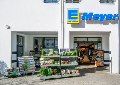 Eingang eines EDEKA-Markts „Meyer“ mit großem Logo; davor Pflanzen- und Warenständer auf dem Gehweg.