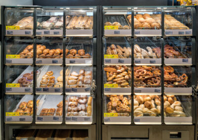 Backwarenregal im Supermarkt mit Brötchen, Brezeln und süßem Gebäck in transparenten Auslagefächern.