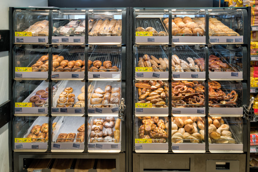 Backwarenregal im Supermarkt mit Brötchen, Brezeln und süßem Gebäck in transparenten Auslagefächern.