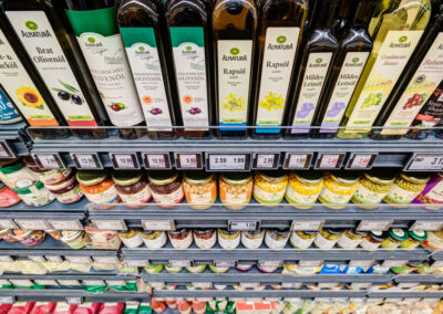 Supermarktregal mit Alnatura-Ölen und Essig (z. B. Olivenöl, Rapsöl, Leinöl) sowie Gläsern darunter; Preisschilder an der Kante.
