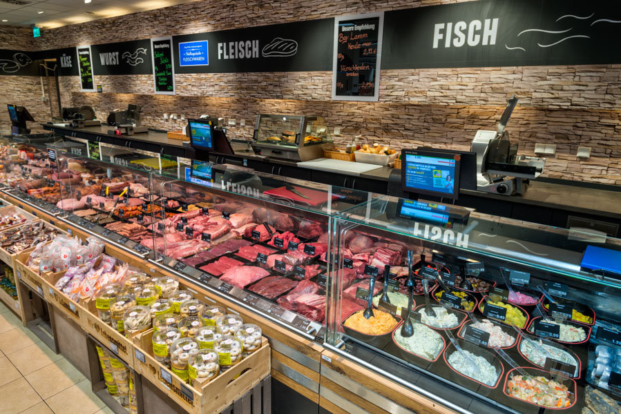 Bedientheke im Supermarkt mit Fleisch- und Feinkostauslage; Beschilderung „Wurst“, „Fleisch“ und „Fisch“ an der Wand.