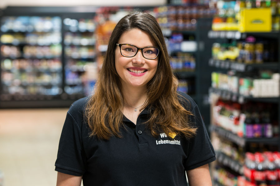 Lächelnde Frau mit Brille in schwarzem Polohemd in einem Supermarktgang; Regale im Hintergrund unscharf.