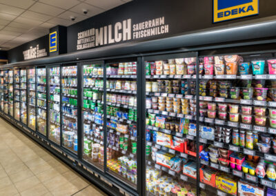 Langes Kühlregal im EDEKA mit Milchprodukten und Desserts hinter Glastüren; darüber große Beschriftung „MILCH“.