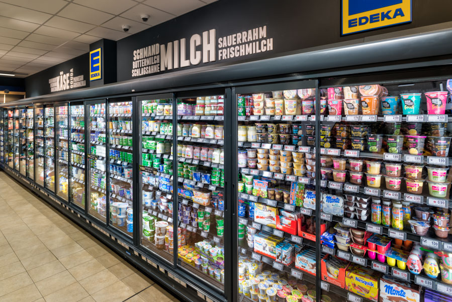 Langes Kühlregal im EDEKA mit Milchprodukten und Desserts hinter Glastüren; darüber große Beschriftung „MILCH“.