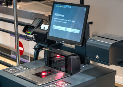 Supermarktregal mit Alnatura-Ölen und Essig (z. B. Olivenöl, Rapsöl, Leinöl) sowie Gläsern darunter; Preisschilder an der Kante. Selbstbedienungskasse im Supermarkt mit Touchscreen („Willkommen“) und Scanner an der Kassenstation.