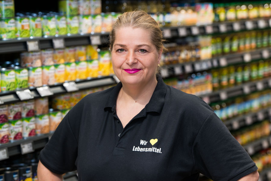 Lächelnde Frau im schwarzen Polohemd mit „Wir lieben Lebensmittel“-Logo vor einem unscharfen Supermarktregal.
