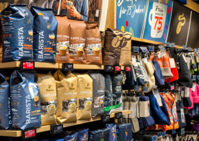 Regal im Supermarkt mit Kaffeebohnen-Packungen (u. a. Barista/Caffè Crema) und daneben ein Ständer mit Mützen und Handschuhen