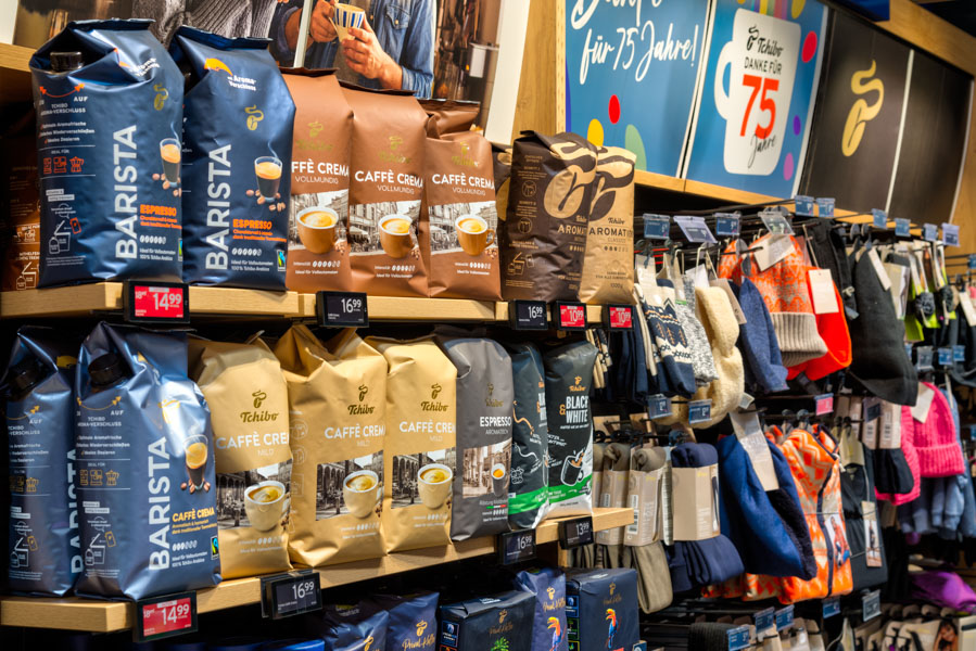 Regal im Supermarkt mit Kaffeebohnen-Packungen (u. a. Barista/Caffè Crema) und daneben ein Ständer mit Mützen und Handschuhen
