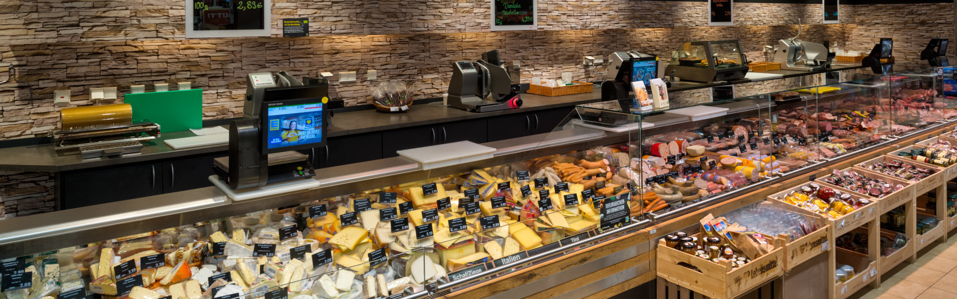 Lange Bedientheke im Supermarkt mit Käse-, Wurst-, Fleisch- und Fischabteilung; beschriftete Tafeln an einer Steinoptik-Wand.