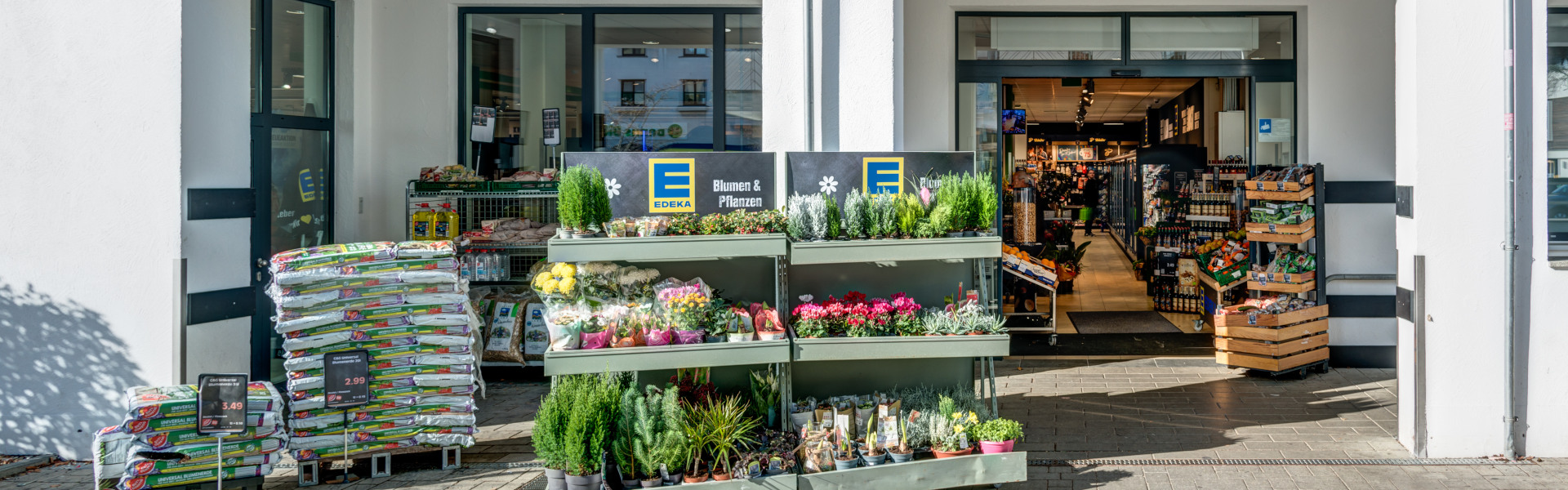 Eingang eines EDEKA-Markts „Meyer“ mit großem Logo; davor Pflanzen- und Warenständer auf dem Gehweg.