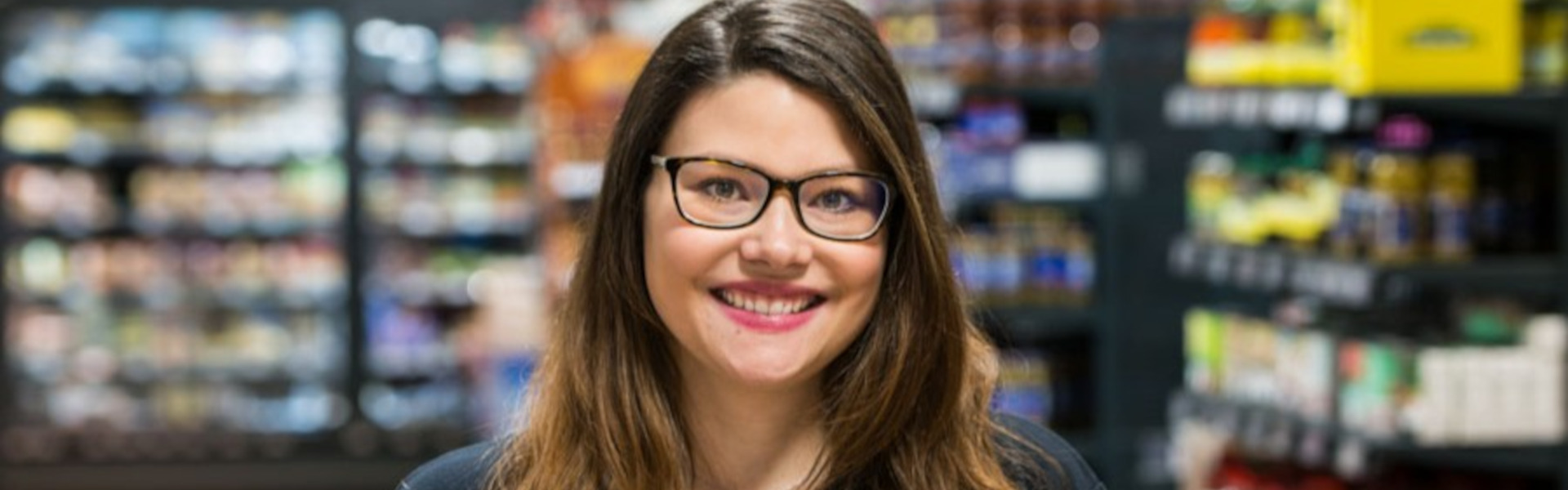 Lächelnde Inhaberin mit Brille in schwarzem Polohemd in einem Supermarktgang; Regale im Hintergrund unscharf.